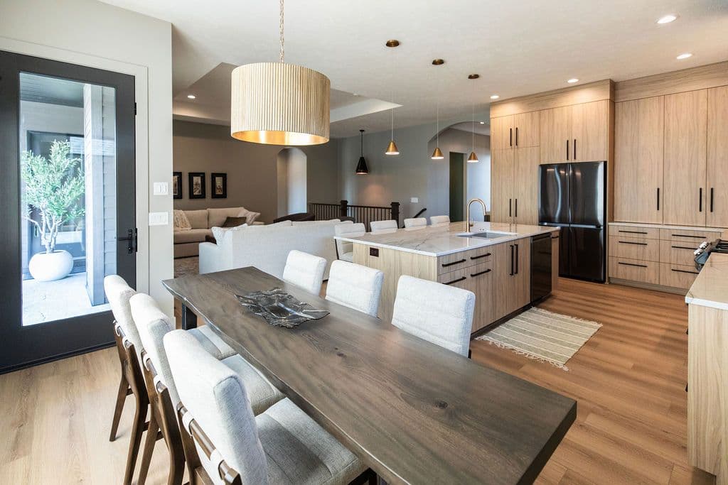 Open-concept dining and kitchen area featuring a long, wooden dining table with upholstered chairs, modern island with marble countertops, and light wood cabinets. A black refrigerator adds contrast, while gold pendant lights and a drum shade chandelier provide illumination.