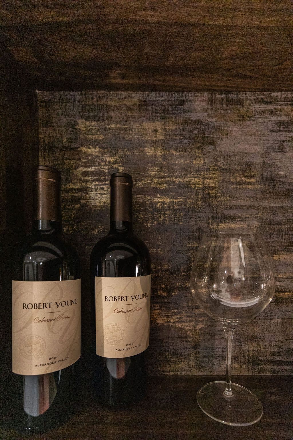 Two bottles of Robert Young Cabernet Franc wine sit next to an empty wine glass against a textured, dark wood background. The wine labels are light beige with dark brown lettering.