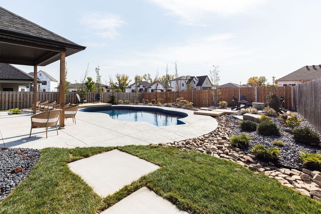 Outdoor space featuring a kidney-shaped pool surrounded by a concrete patio and landscaping. The backyard includes a covered patio area with seating and decorative rock features.