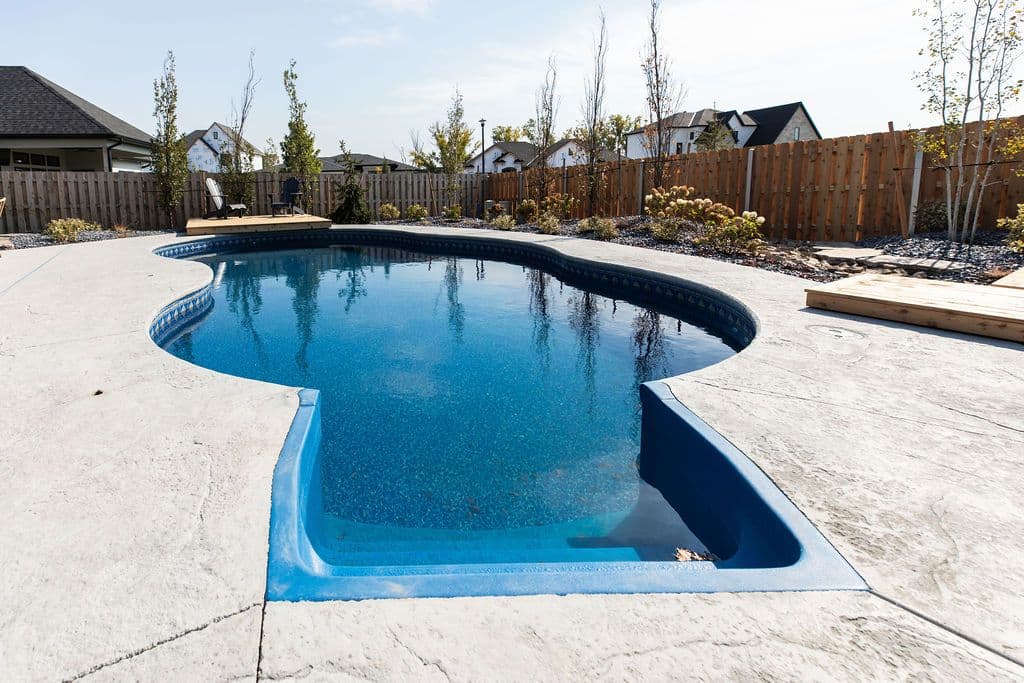 Outdoor pool area featuring a blue pool with concrete surround, wooden deck, and wooden fence. Houses are visible in the background.
