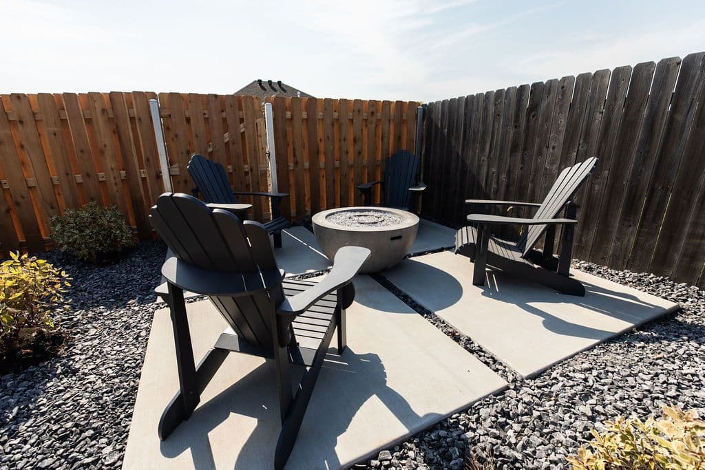 Outdoor seating area with four black Adirondack chairs arranged around a gray concrete fire pit. The area features gray gravel ground cover and square concrete patio stones, enclosed by a wood fence.