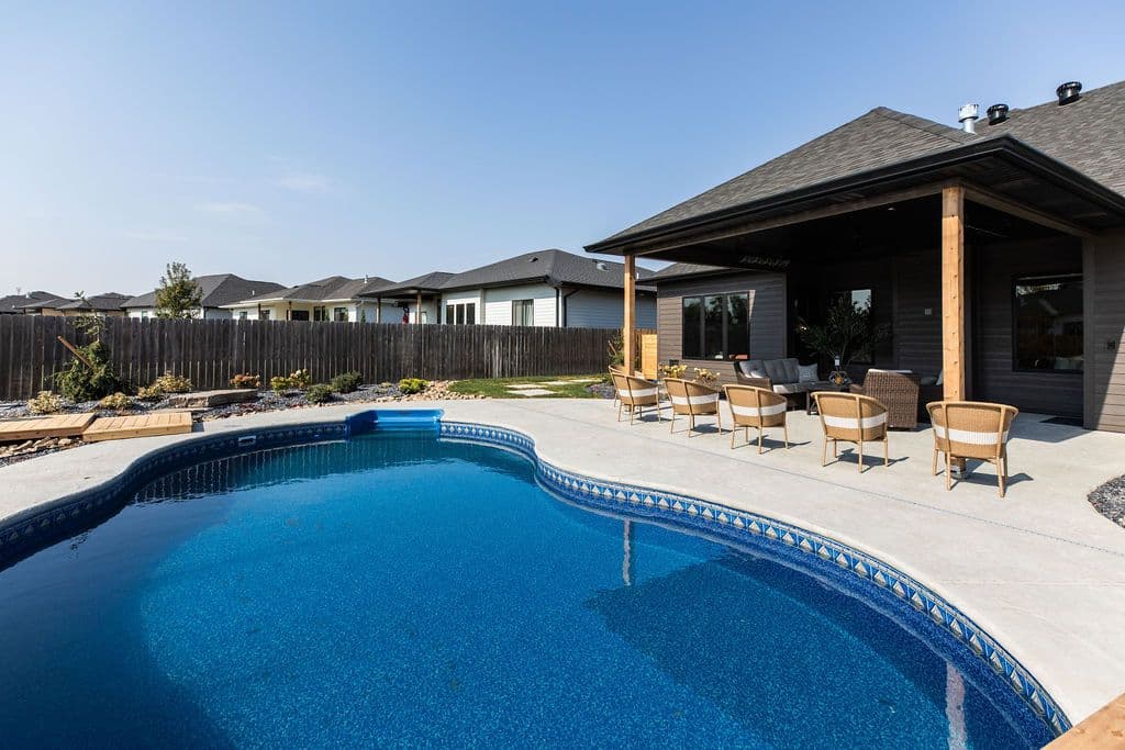 Outdoor patio with a swimming pool and wicker furniture, surrounded by a dark wooden fence. The patio features a covered seating area with views of neighboring homes.