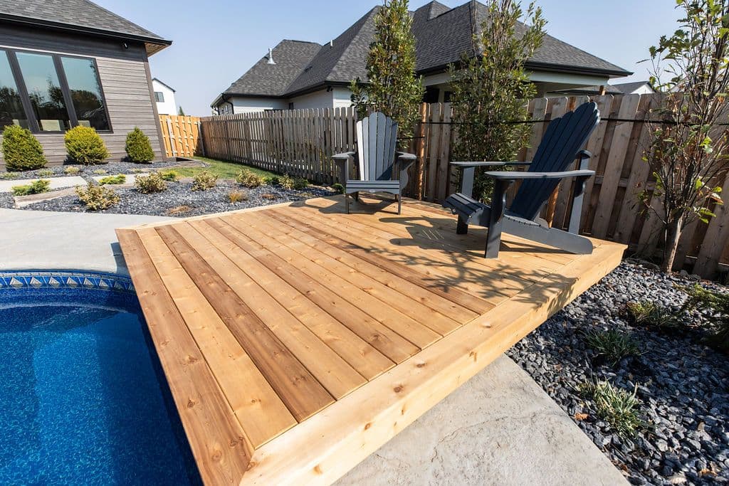 Outdoor deck area featuring wood decking adjacent to a pool, furnished with two Adirondack chairs and surrounded by landscaping and a wooden fence. The scene showcases a backyard setting with residential architecture in the background.