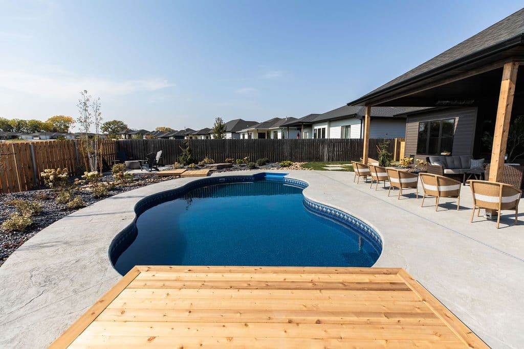 Backyard featuring kidney-shaped pool with wooden deck surrounded by patio and modern outdoor seating. Wooden fence encloses the space with several houses visible in the background.