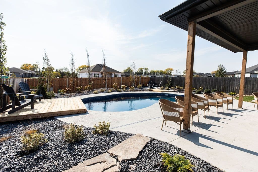 Backyard pool area featuring a kidney-shaped swimming pool surrounded by concrete pavers and black gravel landscaping. A wooden deck with two Adirondack chairs sits to the left, while a covered patio with rattan chairs extends to the right.