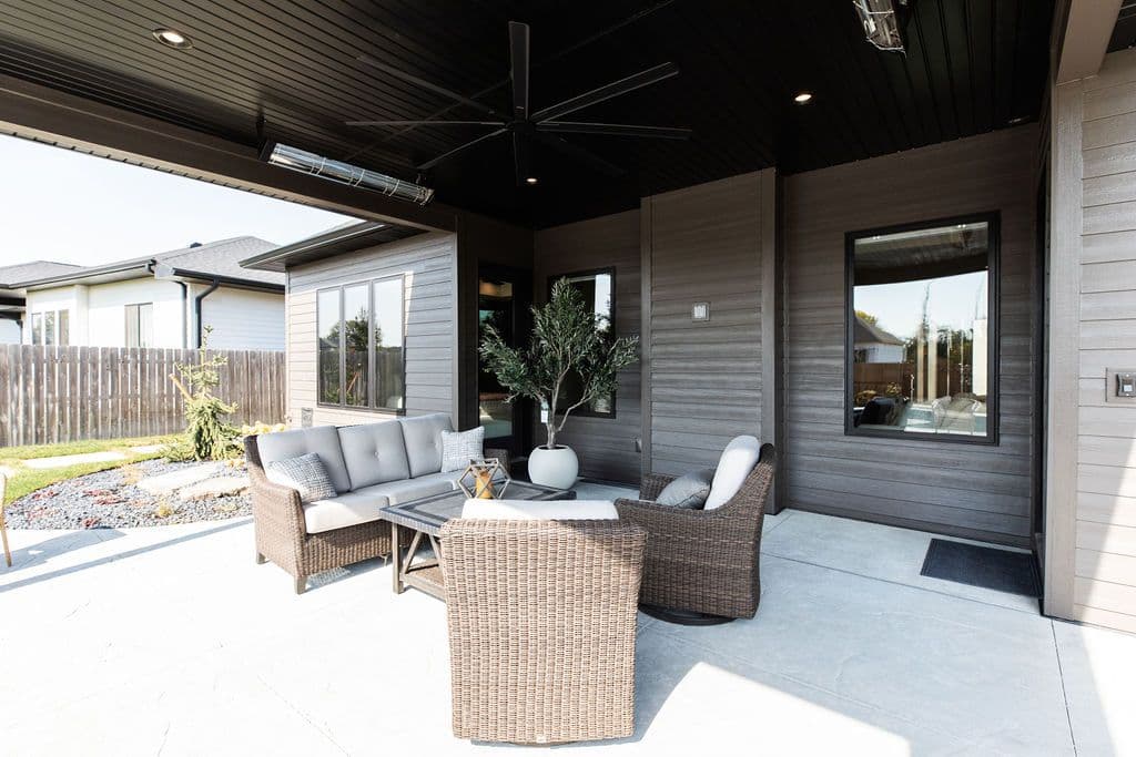 Outdoor patio space featuring wicker furniture set with gray cushions, centered around a low coffee table. The area is covered by a dark ceiling with a ceiling fan and outdoor heater, with large windows and a potted tree visible.