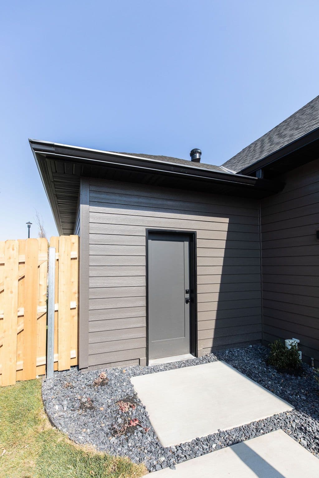 Exterior shot of a home with a grey door, wood-look siding, and a small concrete patio surrounded by grey rocks. There is a partial view of a wooden fence on the left.