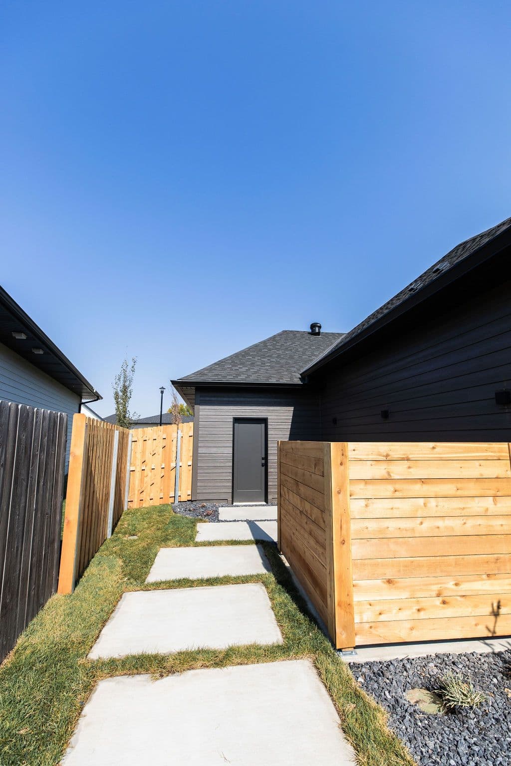 Exterior view features a concrete stepping stone path leading to a gray-stained building with a matching gray door. The walkway is bordered by a combination of light-toned wood and dark-stained wood fencing, with green grass and dark gravel landscaping visible.