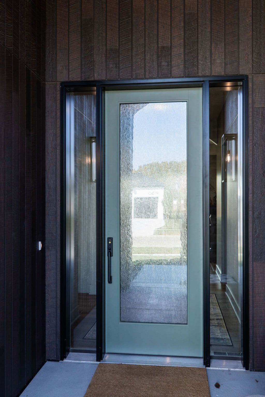 Modern entryway with a sage green front door featuring textured glass, flanked by sidelight windows. The door is set within a dark, wood-paneled facade and leads onto a concrete porch with a small doormat.