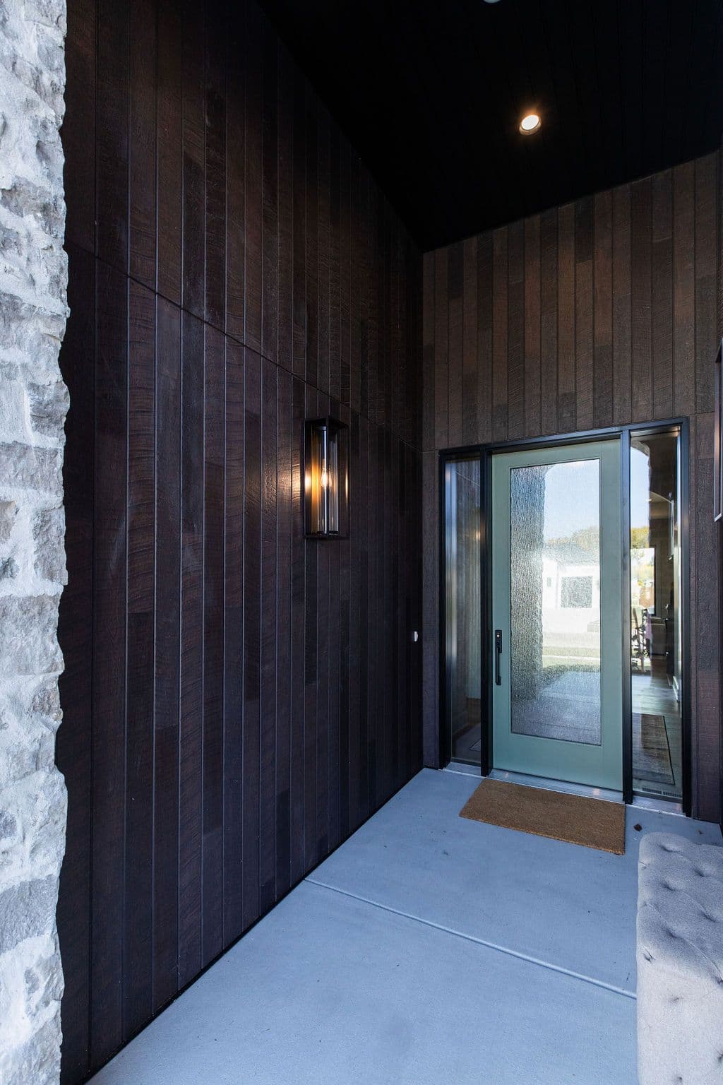 Exterior view showcases a modern entryway with a concrete patio leading to a sage green framed door, accented by a textured glass panel. The surrounding wall features a rich dark brown wood paneling, complemented by a stone veneer section on the left and an outdoor light fixture.
