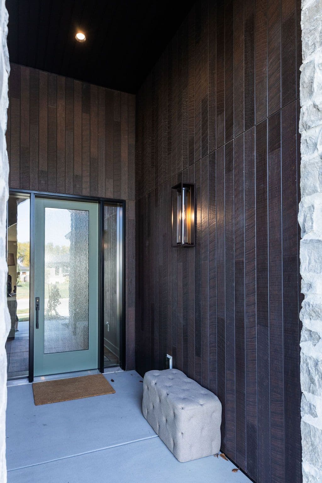 Exterior entryway featuring a dark wood paneled wall with a modern light fixture and a light grey upholstered bench. The entryway leads to a front door with textured glass, and the space is framed by stone cladding.