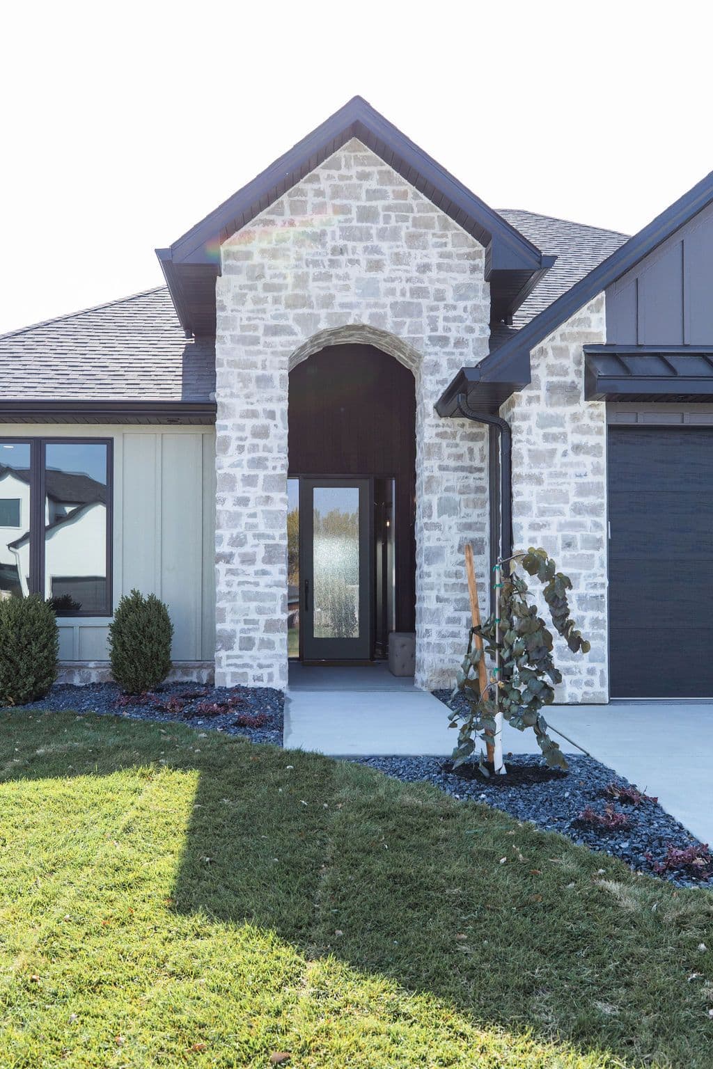 Exterior view of a modern home featuring a stone veneer archway entrance, contrasting gray horizontal siding, and dark gray garage door. Landscaped with green grass, small bushes, and dark gravel.