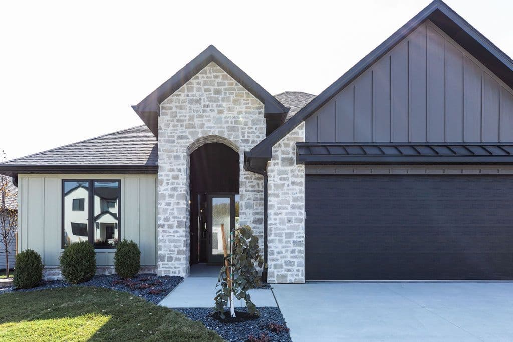 Home exterior featuring stone veneer entry, dark garage door, and light siding. A walkway leads to the front door, and there are shrubs and landscaping.