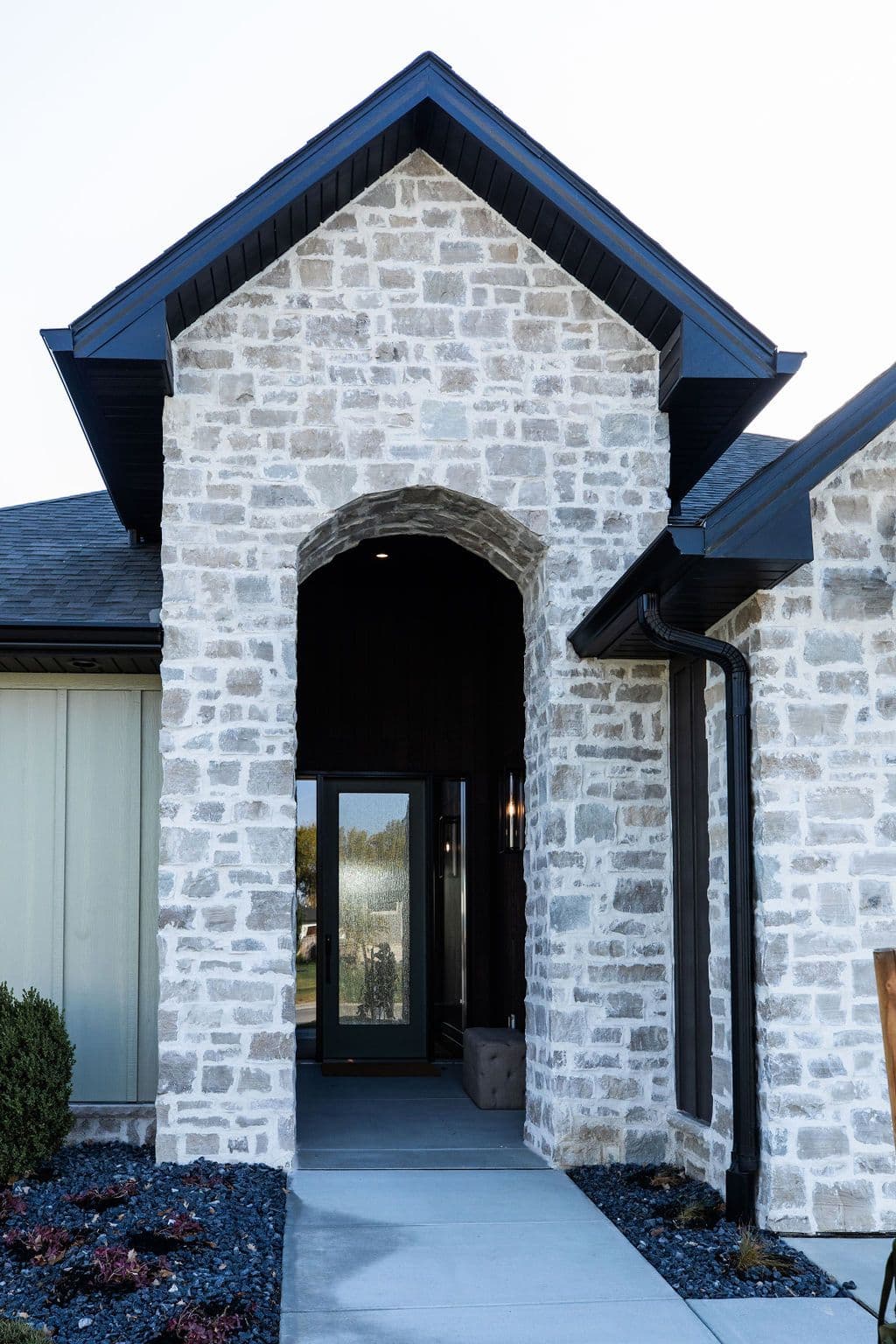 Stone facade entrance with arched doorway and black trim and accents. Concrete walkway leading to glass front door.