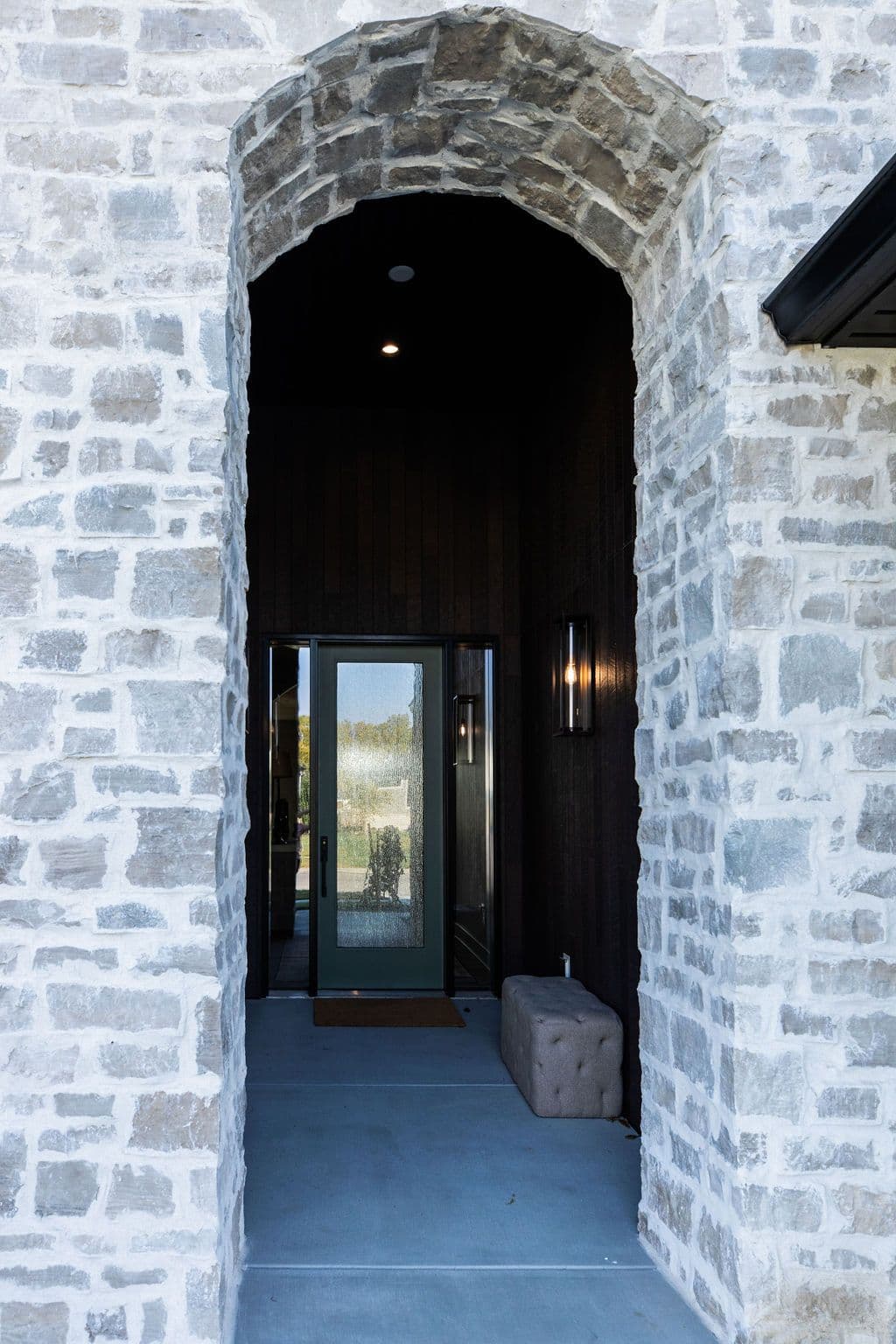 Exterior entrance to a home featuring a stone archway framing a dark wood-paneled entryway with a glass door. Modern exterior lighting and concrete flooring complete the sophisticated look.