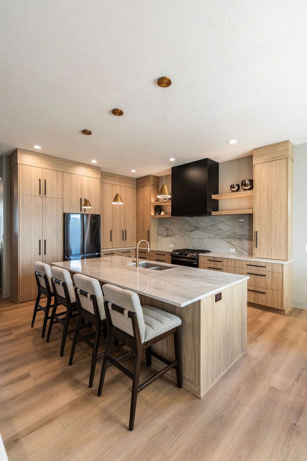 Modern kitchen with light wood cabinets, marble countertops, and island seating. Pendant lighting hangs above the island and stainless steel appliances are visible.