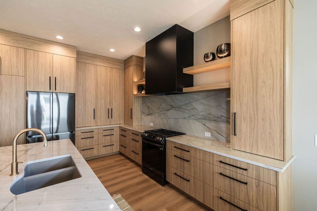 Modern kitchen with wood cabinetry, black hardware, and marble countertops. A black range hood is centered over a black range, and a stainless steel refrigerator is integrated into the cabinetry.