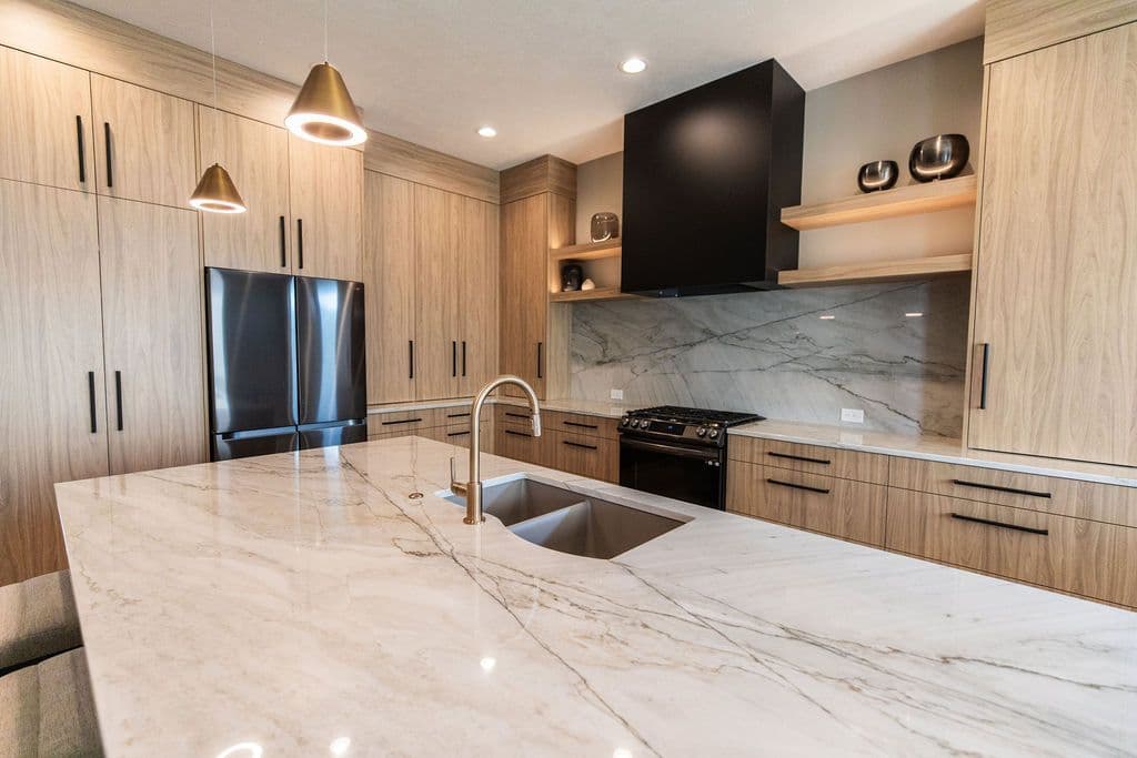 Modern kitchen featuring light wood cabinetry with black hardware and a large marble island with a built-in sink and brass faucet. A black range hood and stainless steel appliances complete the contemporary design.