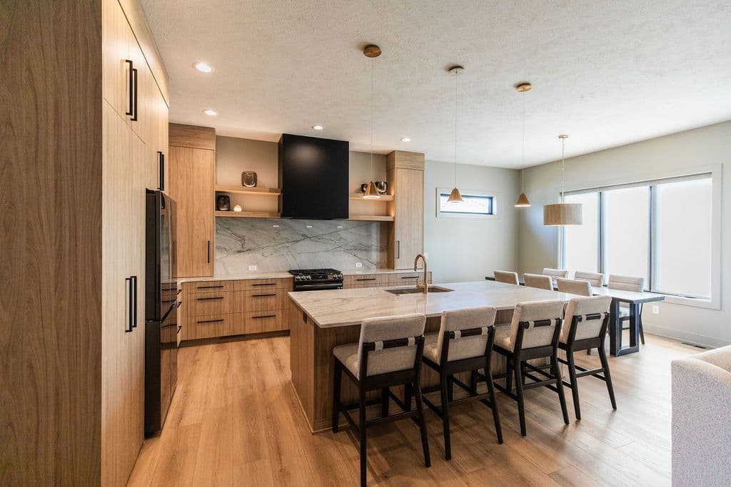 Modern kitchen with light wood cabinetry, black hardware, and a large island with seating. The kitchen features marble countertops and backsplash, a black range hood, and pendant lighting above the island and dining area.