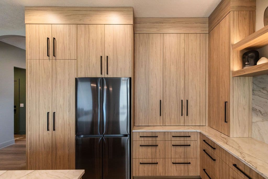 Modern kitchen features light wood grain cabinetry with sleek black hardware and stainless steel appliances. The space is illuminated by warm recessed lighting and includes open shelving with decorative objects.