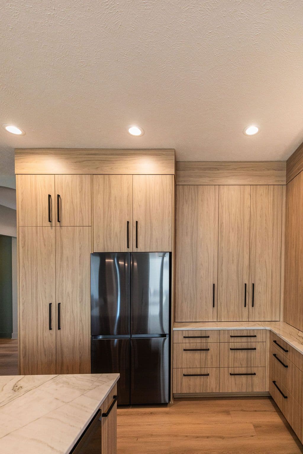 Modern kitchen featuring light wood cabinetry with sleek black hardware and stainless steel refrigerator. White marble countertops are visible on the island and surrounding cabinets.