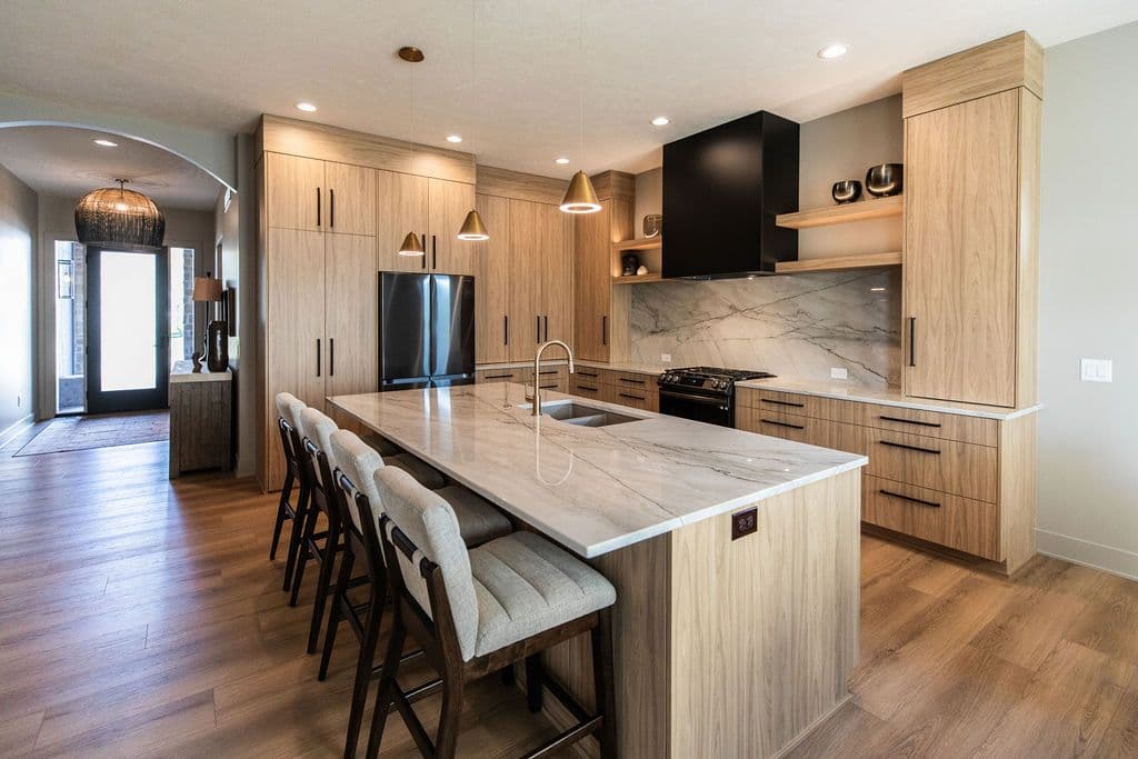 Modern kitchen with light wood cabinetry, marble countertops, and a black range hood. There is a large island with seating and warm lighting fixtures.