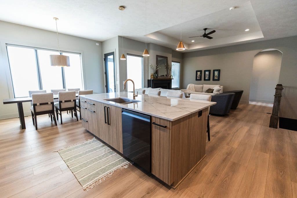 Bright, modern open concept kitchen and living area features a large island with sink, pendant lighting, and wood-look cabinetry, a dining area with large windows, and a living room with recessed lighting and fireplace.
