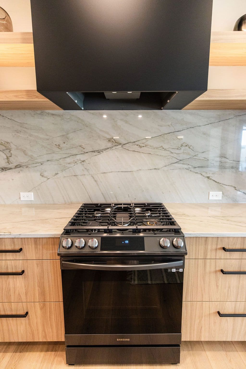 Modern kitchen featuring a black stainless steel gas range with matching range hood above. The kitchen also has light wood cabinets with black hardware, and a light countertop with a marble backsplash.