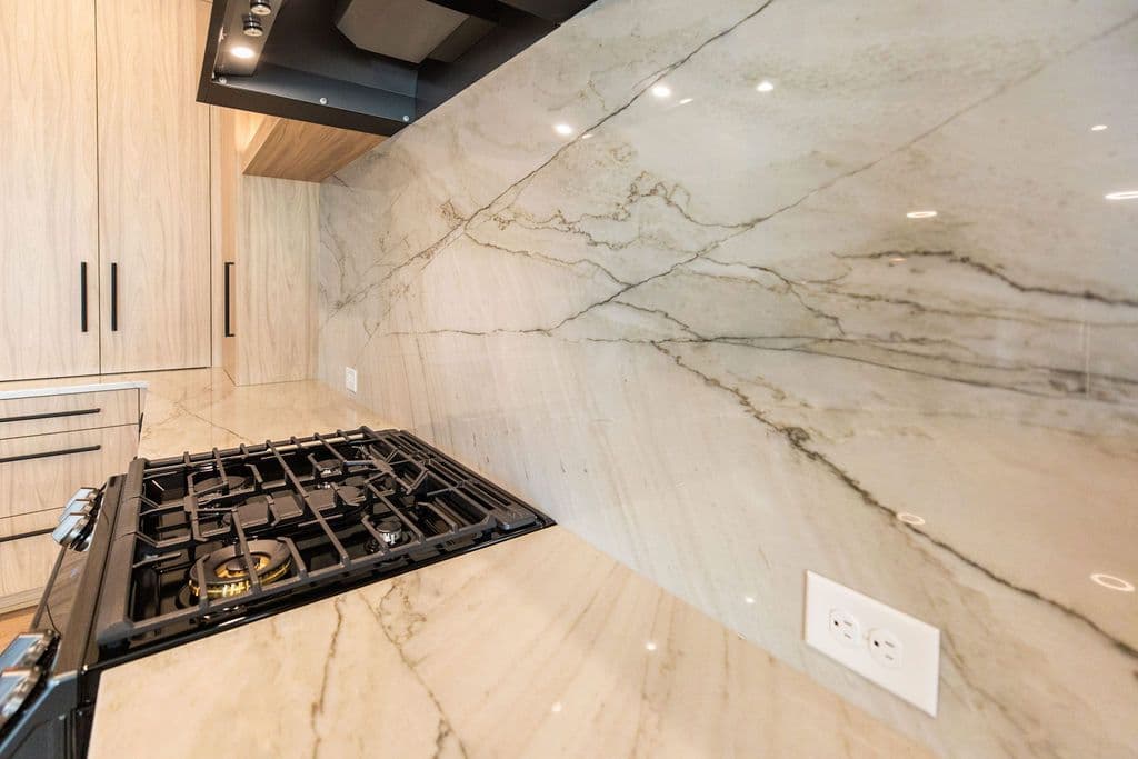 Modern kitchen featuring slab cabinetry, light countertops with marble-like veining extending up the wall as a backsplash, and black gas range. An electrical outlet is visible on the counter.