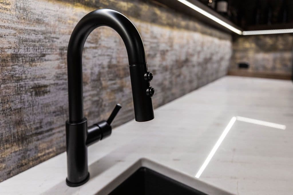 Modern kitchen features a sleek matte black faucet with a pull-down sprayer and buttons, set against a faux-stone backsplash. The white countertop with integrated LED lighting and a black sink create a clean, contemporary aesthetic.