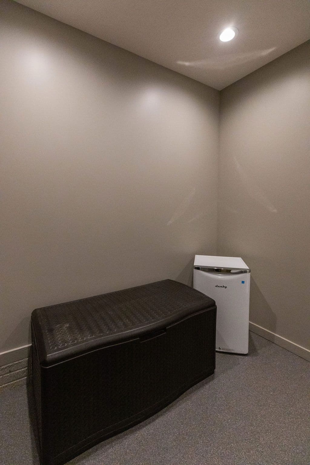 This is a storage room featuring a large brown storage bin and a small white refrigerator. The walls are a neutral tan color, and the floor is covered with gray carpeting.