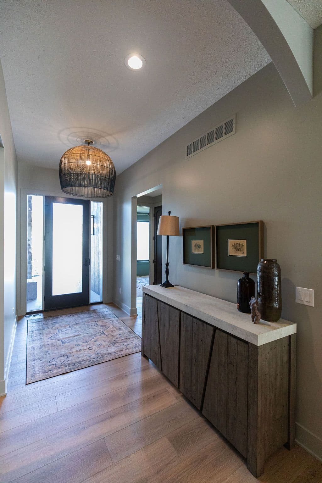 Entryway featuring a wood console table topped with marble, two framed pieces of art, and decorative vases. The space is illuminated by a modern pendant light and a large rug adds pattern to the entryway.