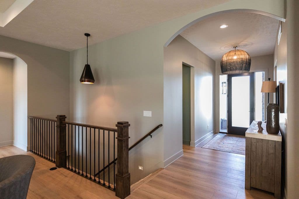 An interior hallway with wood floors and an archway that leads to the front door. A dark metal pendant light hangs in the hallway, and a wooden railing and spindles line the staircase.