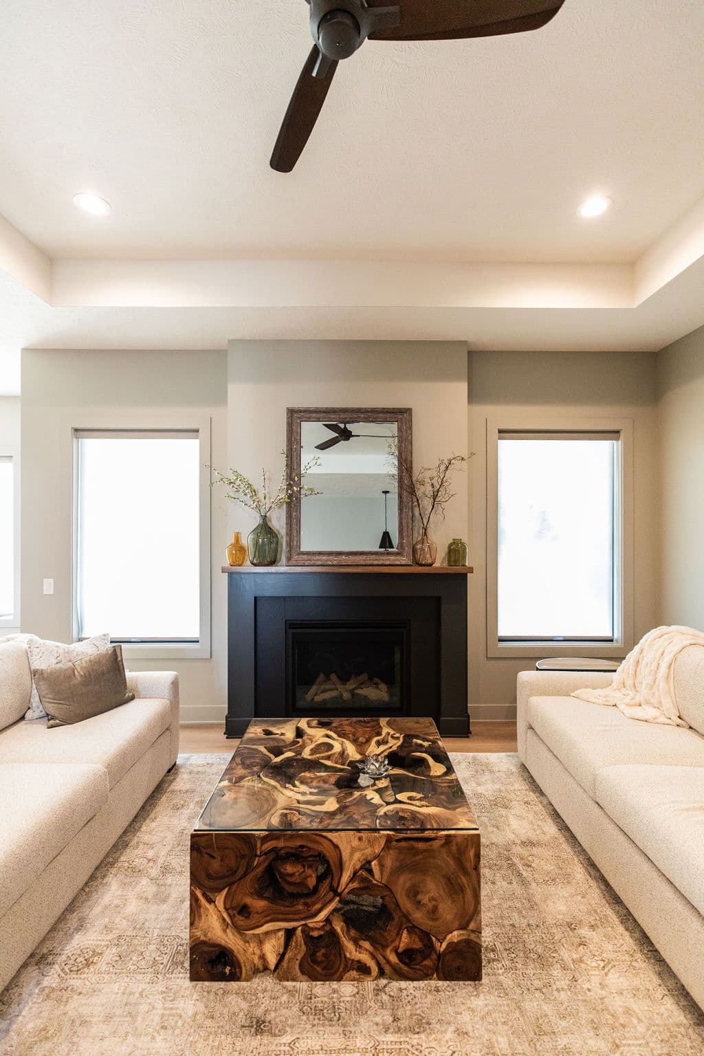 Living room with neutral color palette features a textured wood coffee table, beige sofas, a dark fireplace, and a framed mirror above the mantle. Windows on either side of the fireplace provide natural light.