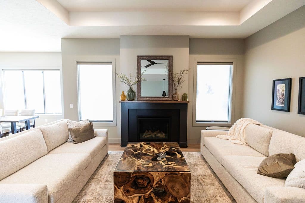 Living room with two cream-colored sofas facing each other across an ornate wood coffee table. A black fireplace with a framed mirror is centered on the wall between two windows.