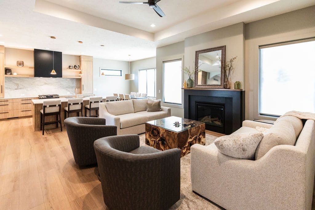 Open concept living room and kitchen featuring hardwood floors, modern cabinets, a black fireplace, and neutral furniture. The kitchen has a large island with bar seating, while the living area features a unique wood and glass coffee table.