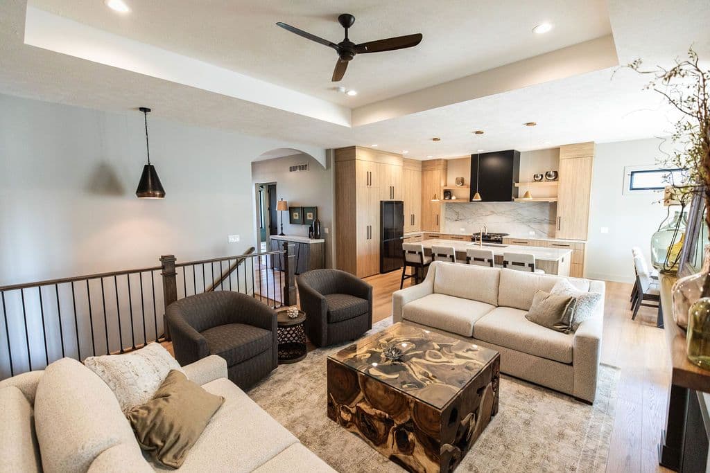 Open concept living room and kitchen with light wood flooring. The living room features two sofas, two armchairs, and a unique wood coffee table.