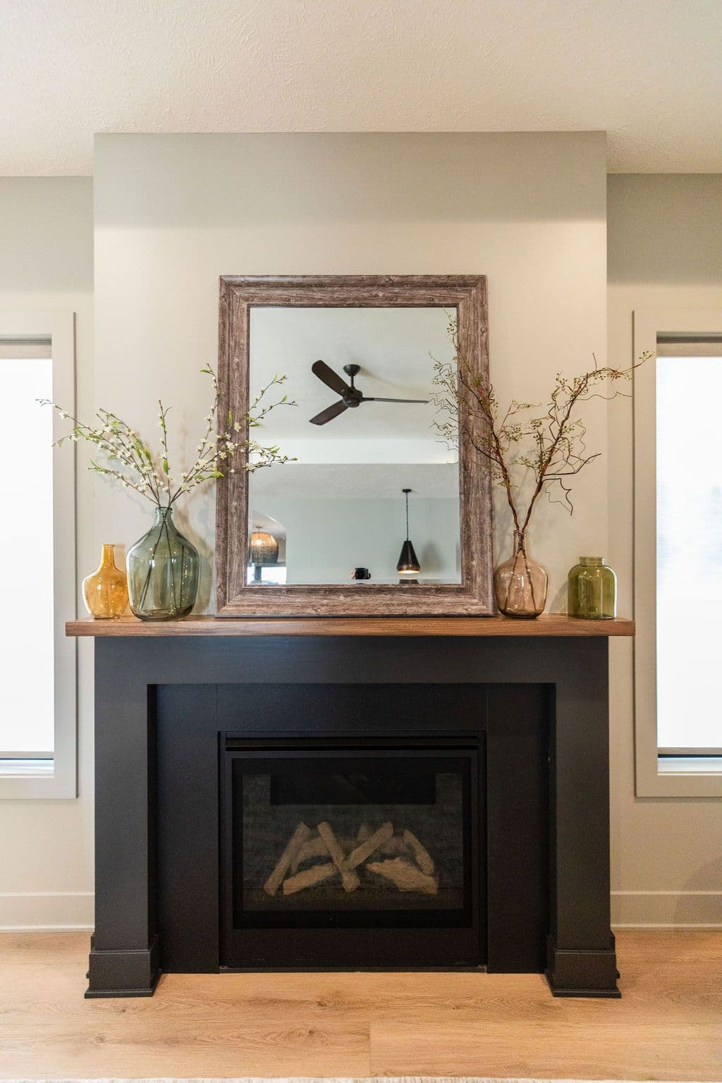 A modern fireplace is framed by a dark mantle with a wooden top, displaying a mirror reflecting a ceiling fan. Three decorative vases sit on the mantle, adding a touch of color and natural elements to the space.