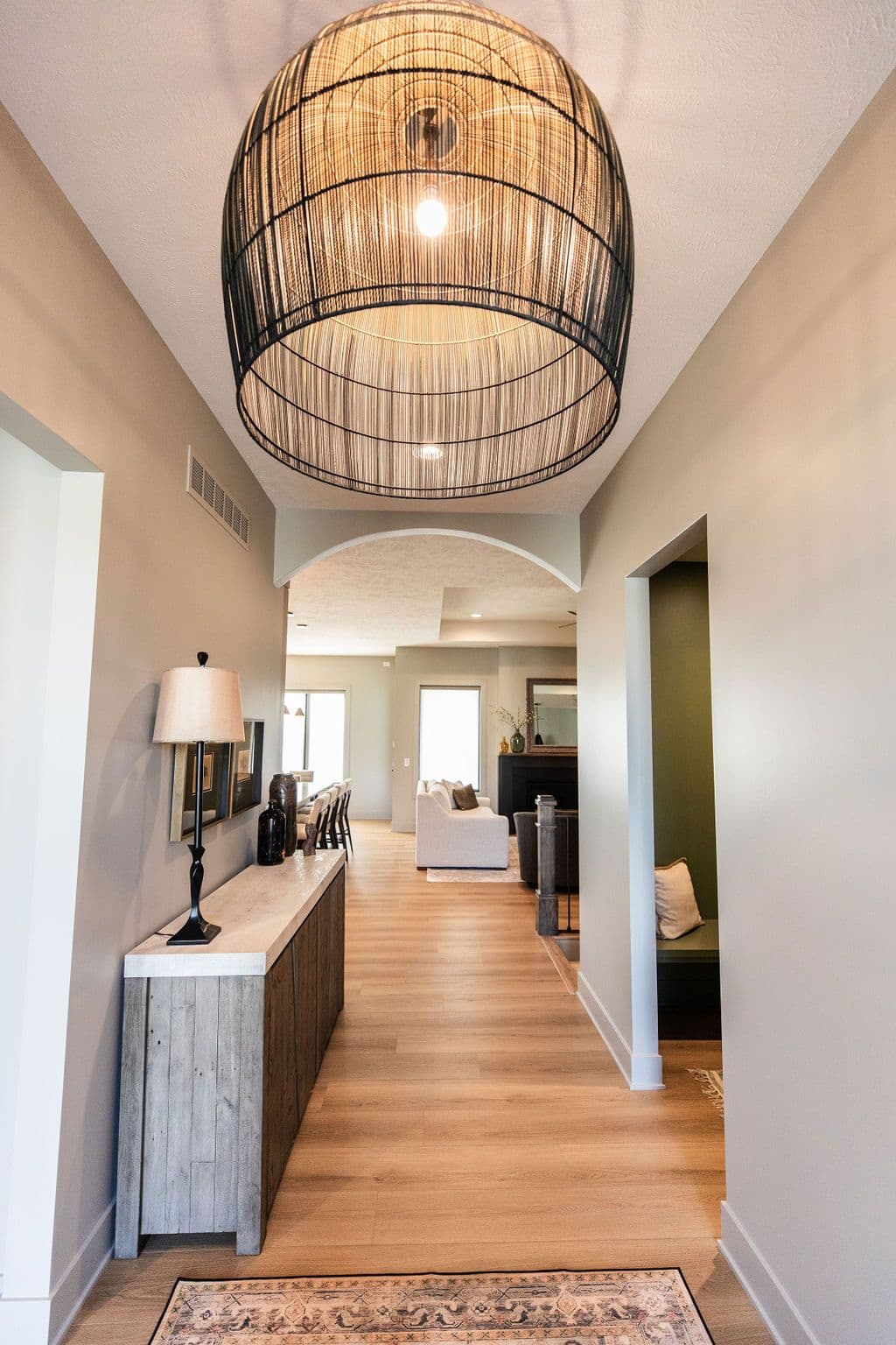 Hallway featuring wood flooring, a large wicker pendant light, a wooden sideboard with a lamp, and an arched doorway leading into the main living space. A small rug is visible at the end of the hallway.