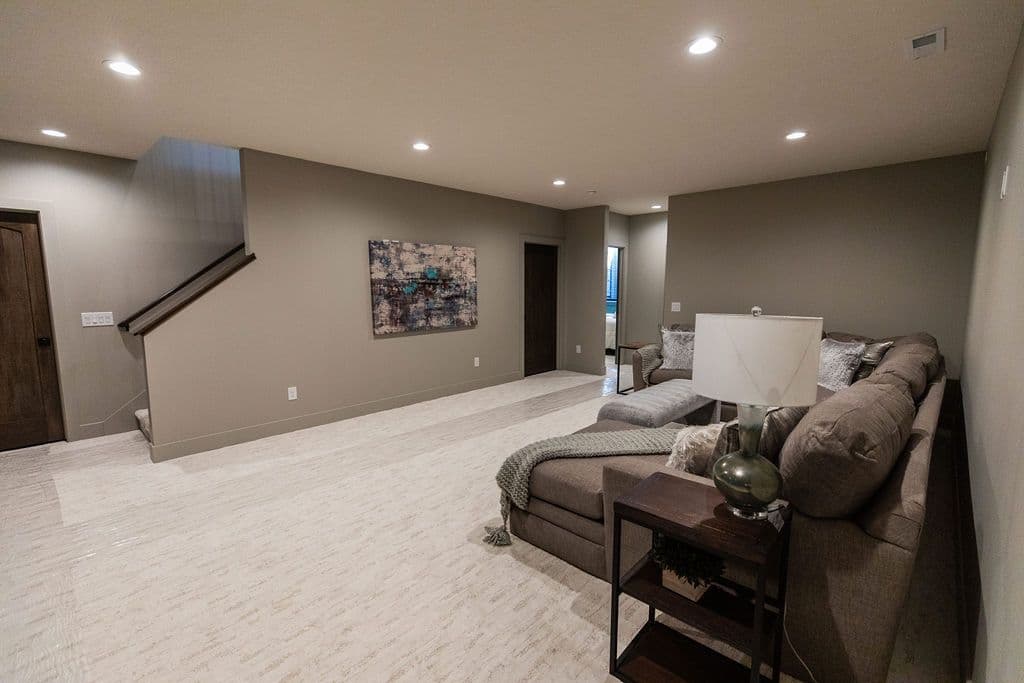 The living room features a gray sectional sofa with a side table, textured off-white flooring, neutral-toned walls, and recessed lighting. The room also includes artwork and a staircase.