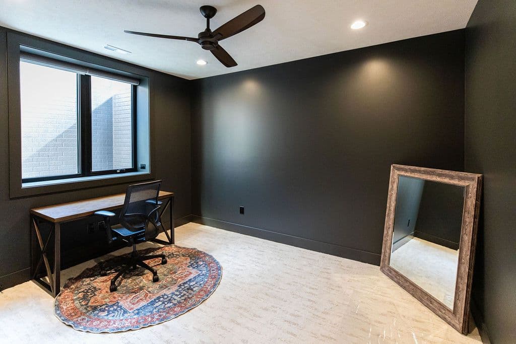 Office space features a dark gray wall, desk and chair situated on an area rug, a mirror leaning against the wall, and a window with a view of a brick wall. A dark brown ceiling fan hangs from the white ceiling.