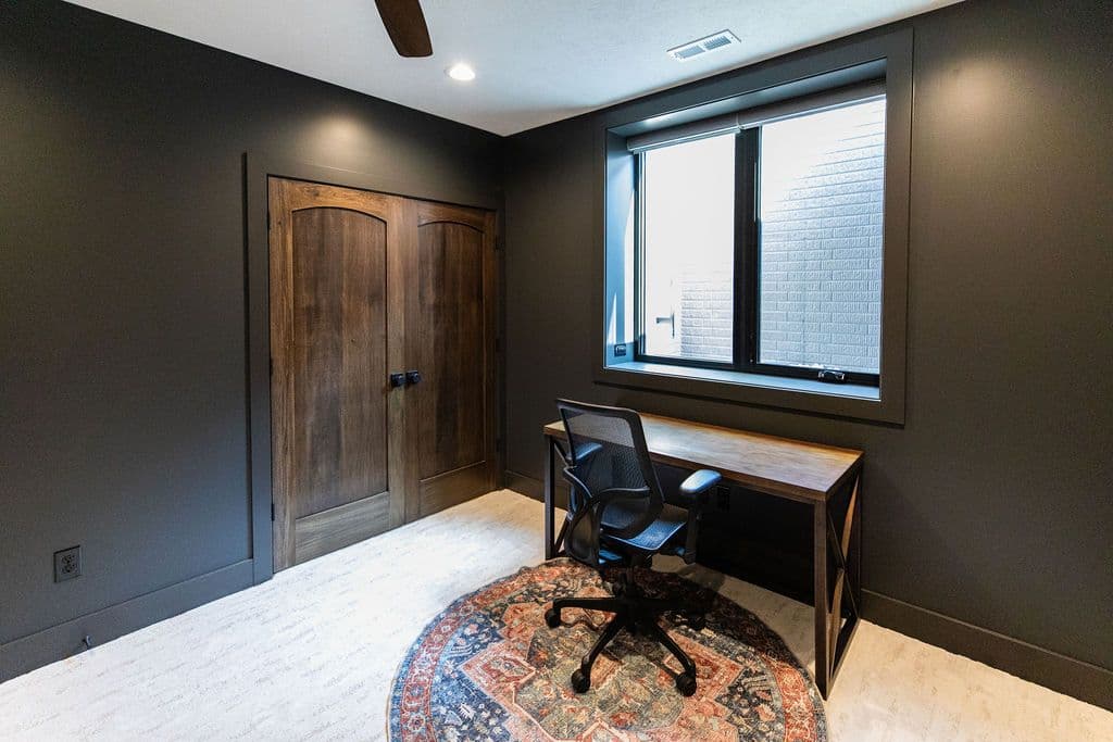 This office features a dark gray color scheme with a rustic wood desk and matching doors. A round patterned rug sits beneath a mesh office chair.