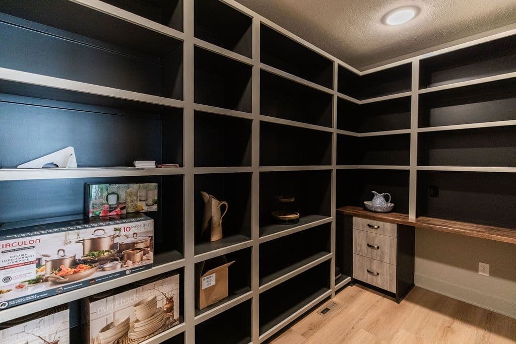 Walk-in pantry featuring extensive built-in shelving painted in contrasting black and white tones, hardwood flooring, and a small countertop area with drawers for storage.