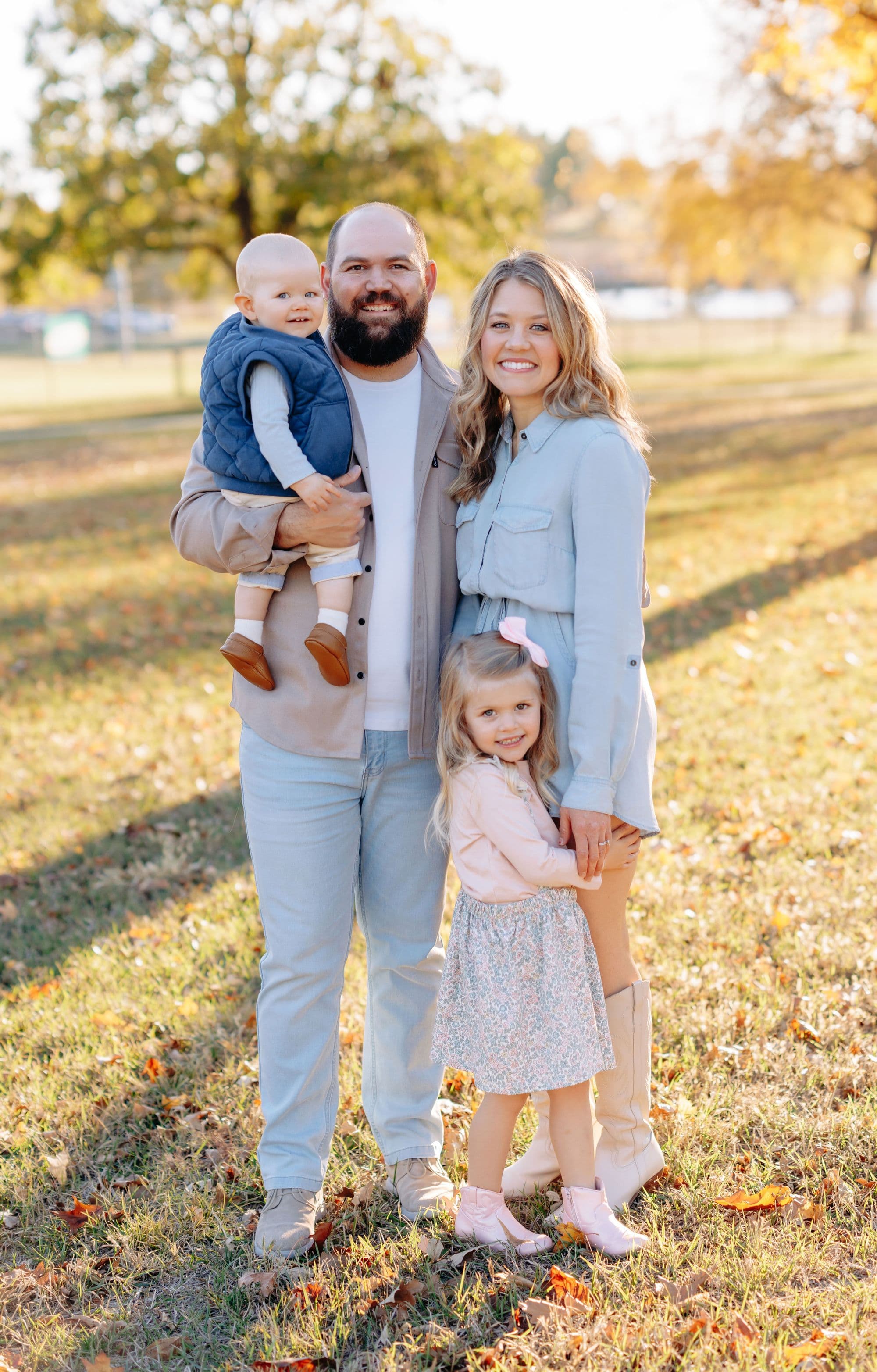 Family portrait features a mother, father, toddler son, and young daughter, posing outdoors on a grassy lawn with autumn foliage. They wear coordinated light-colored outfits in shades of denim and beige.