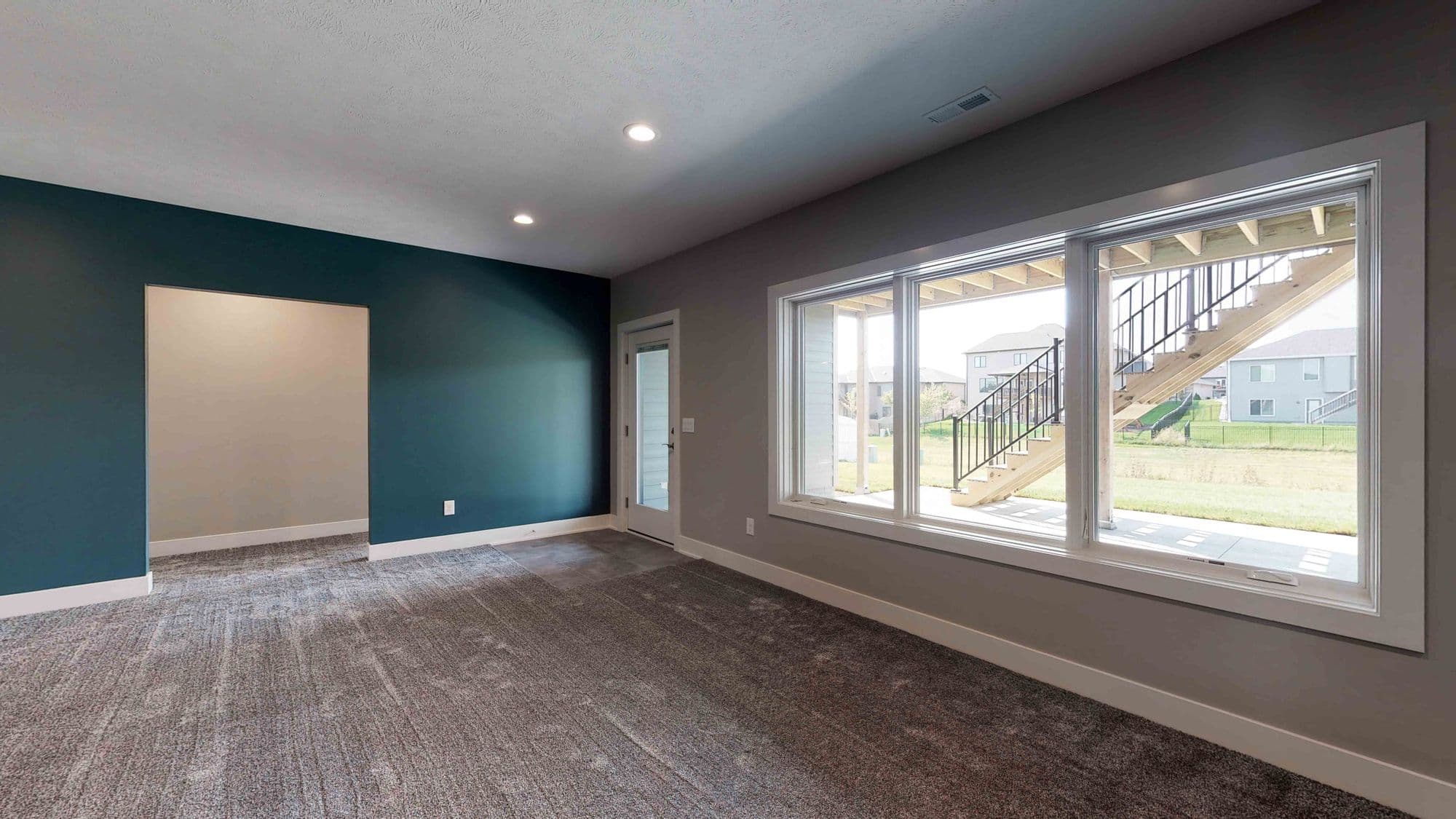 Interior basement room view showing dark teal accent wall, neutral carpeting, and large windows overlooking outdoor stairs. White trim contrasts against the soft gray walls.