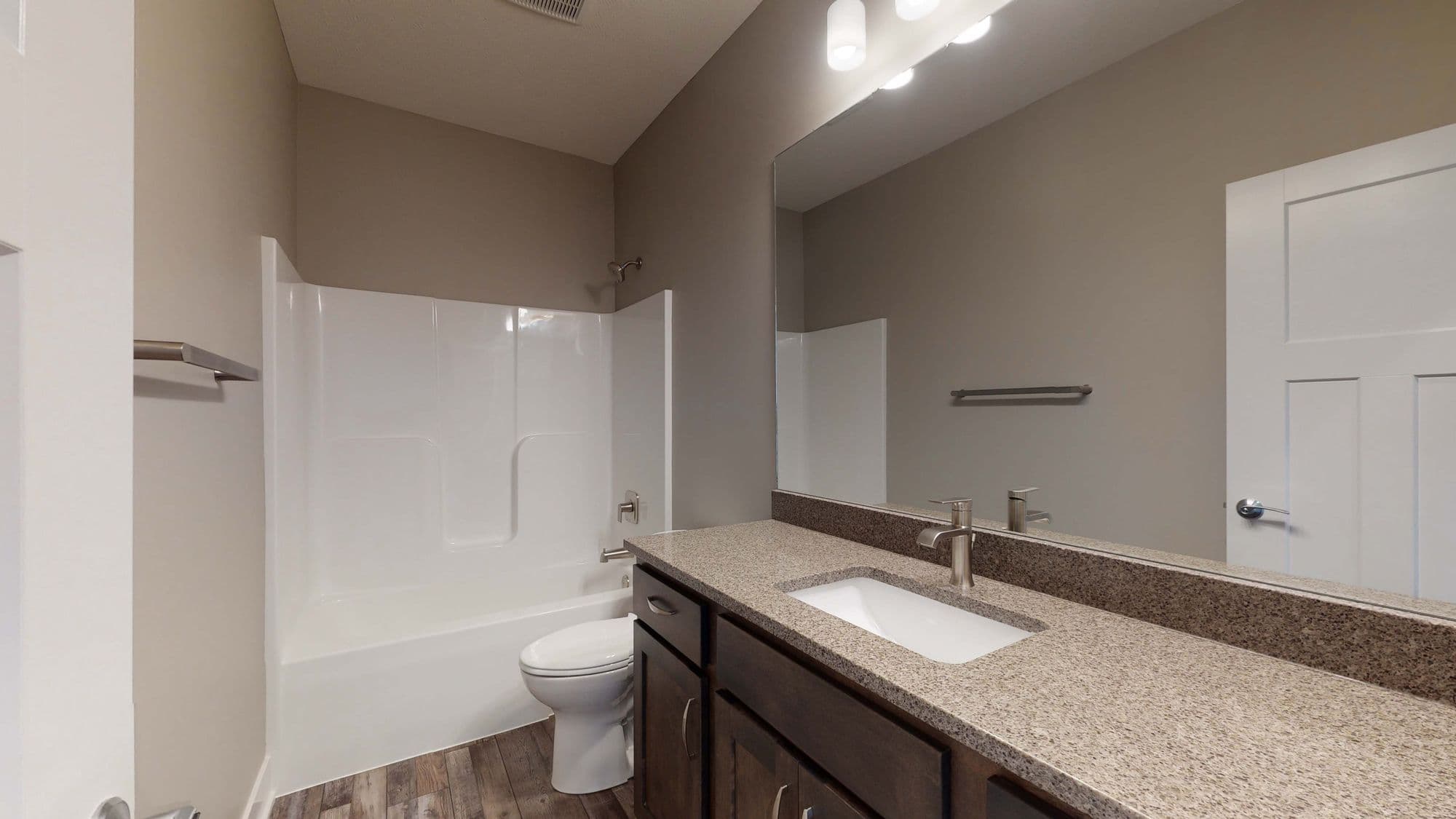 Bathroom featuring a white tub/shower combination, a granite countertop with a white sink, and dark brown cabinets. The room has a large mirror and beige walls.