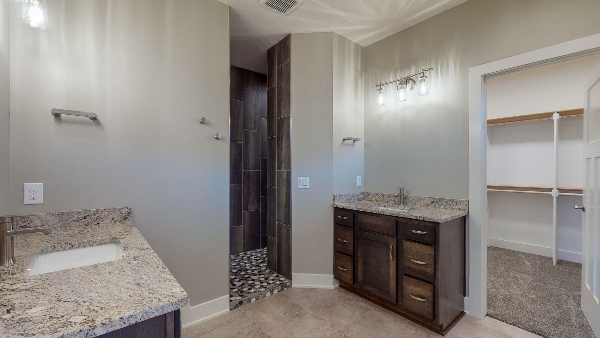 Modern bathroom featuring double vanity with granite countertops, dark wood cabinets, and a tiled shower with mosaic floor. The room includes a walk-in closet with shelving.