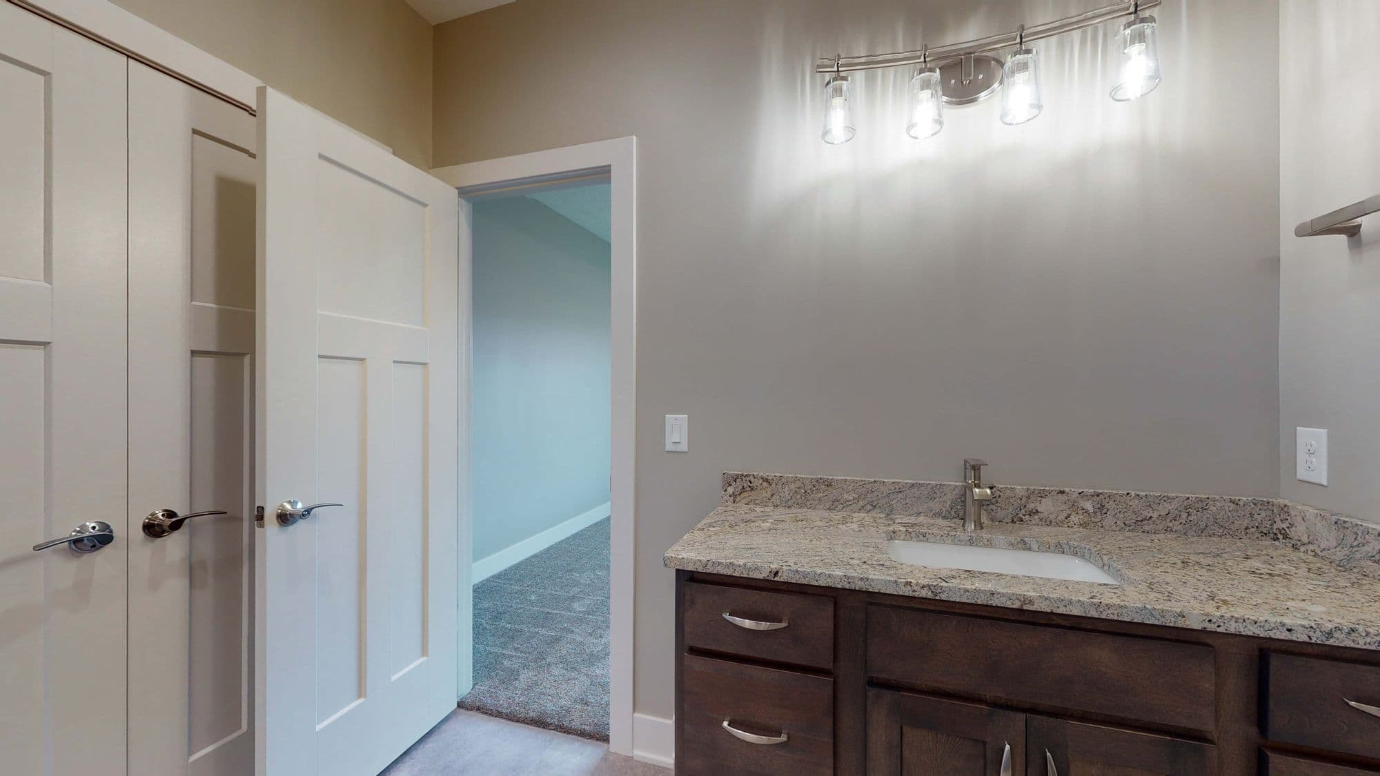 Bathroom interior with dark wood vanity topped with granite, bright vanity lighting, and white paneled doors. An open doorway leads to another room with neutral carpet.