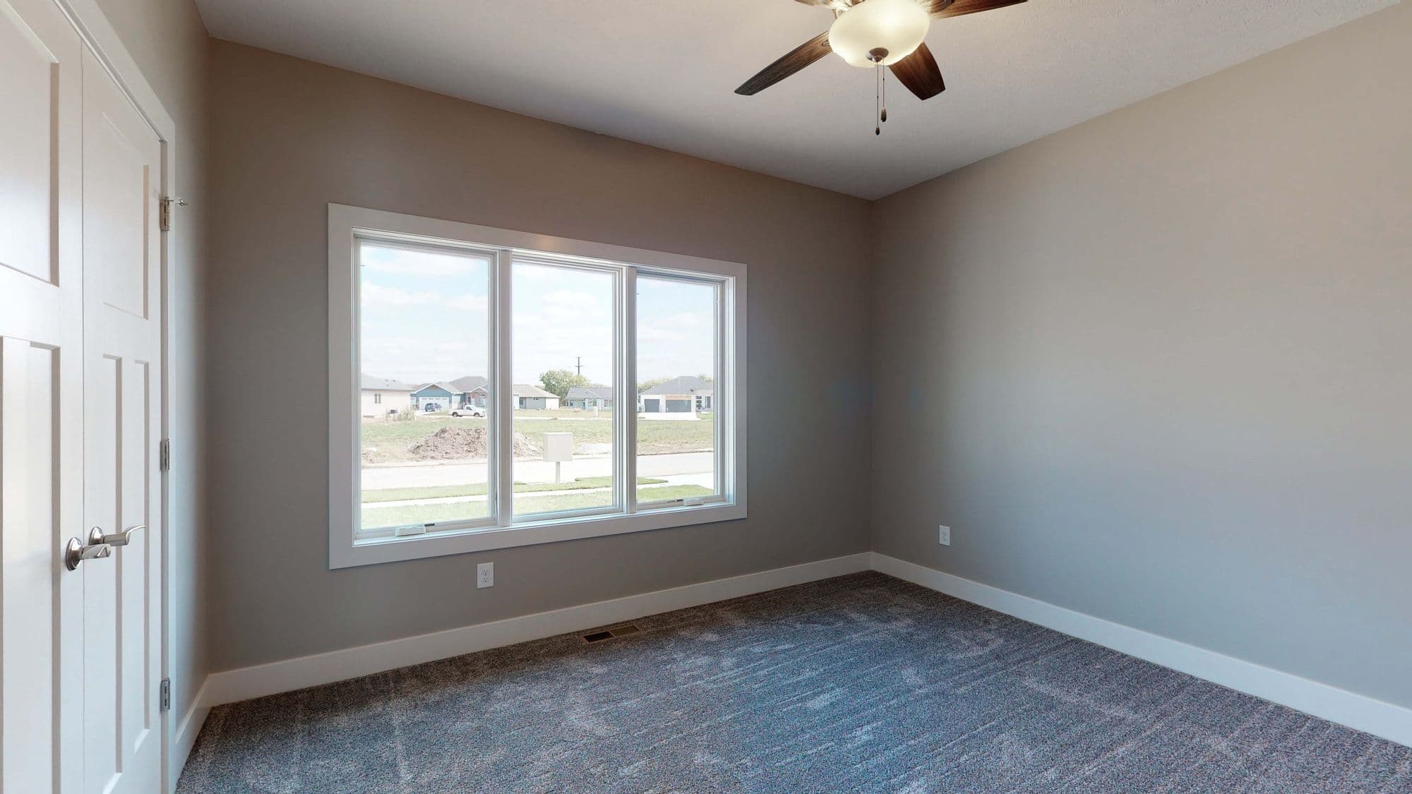 Bedroom featuring a large window with natural light, neutral gray walls, and gray carpeting. A white ceiling fan is visible, along with white trim and a paneled white door.
