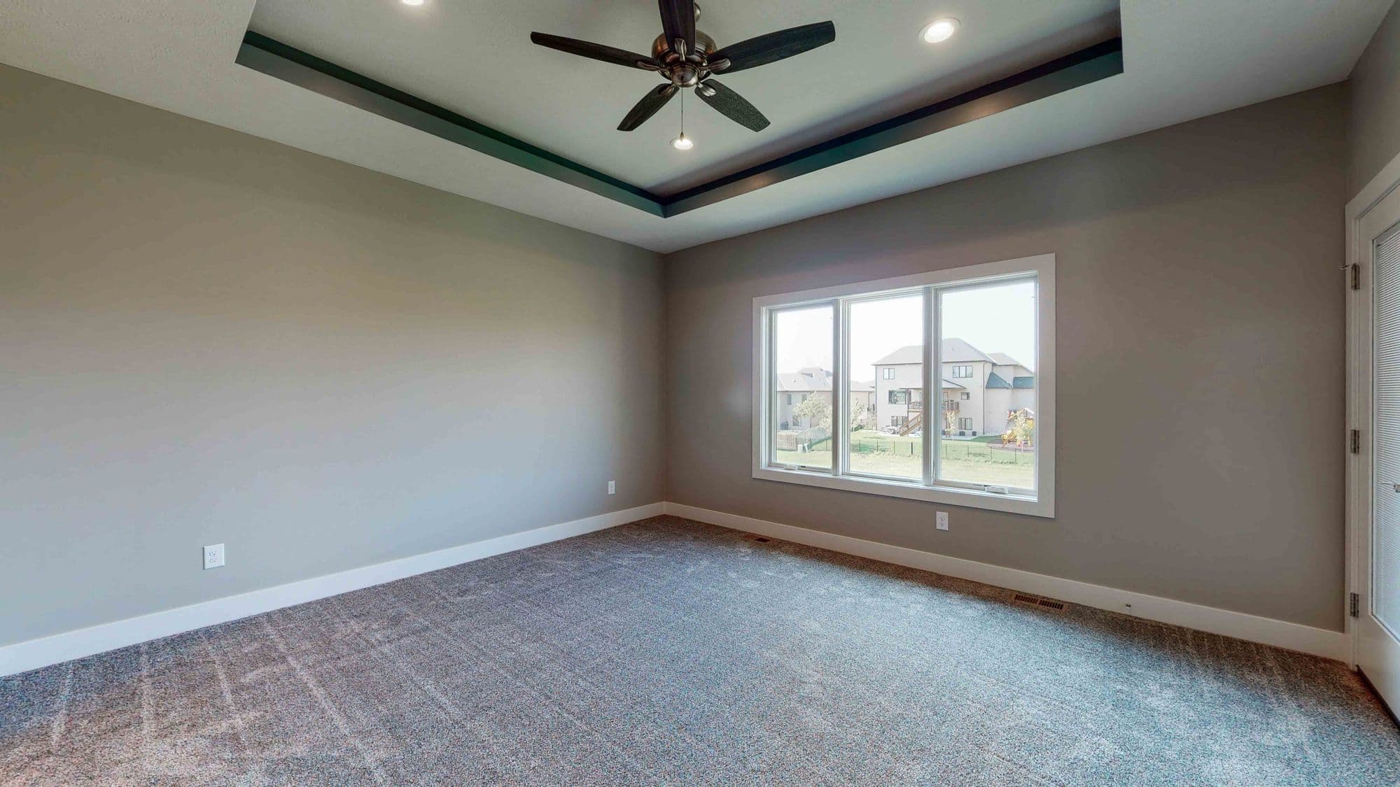 A spacious bedroom features a tray ceiling with a dark accent, a large window overlooking a residential area, and neutral-toned walls. The room is carpeted with a gray patterned carpet and has white trim.